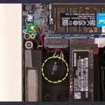 A laptop motherboard with the round CMOS coin cell battery circled in yellow, beside RAM and an M.2 SSD